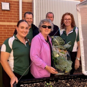 Group of five adults smiling beside a raised garden bed, holding freshly harvested greens, with a brick wall and white fence in the background.